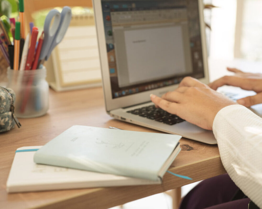 Schreiben mit Gefuehl_Julia Hofmarcher Zwei Hände tippen in einen Laptop, links neben den Händen liegen zwei kleine Bücher.