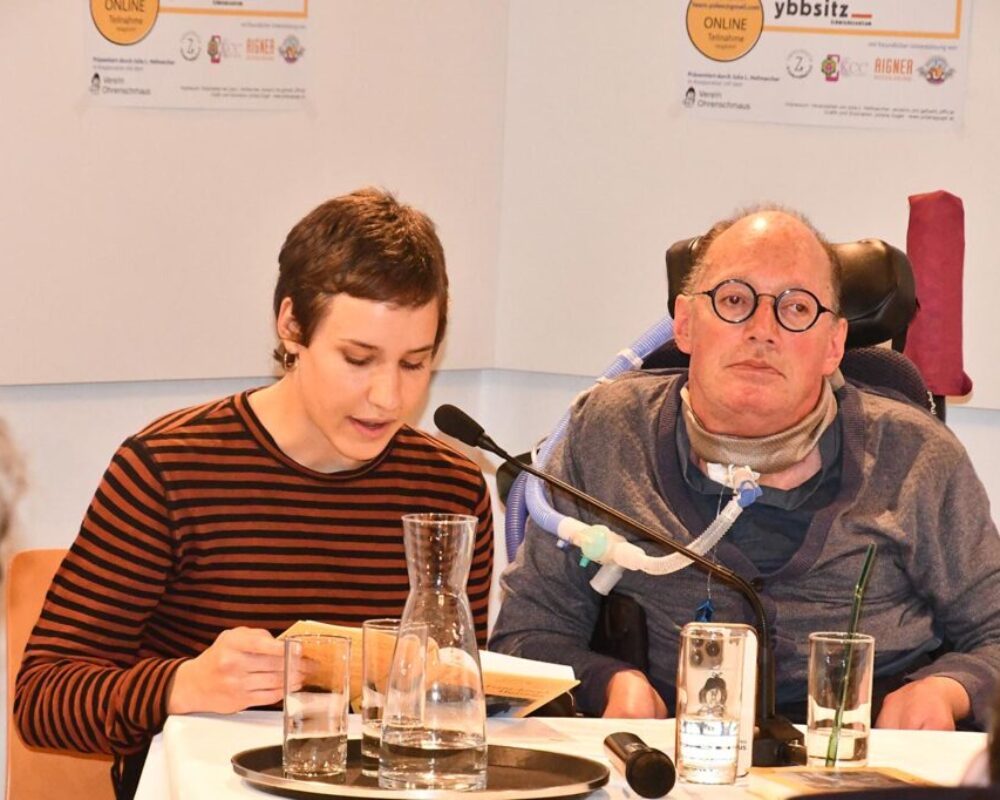 Huainiggs Persönliche Assistentin liest aus seinem Buch. Er sitzt neben ihr und hört zu.