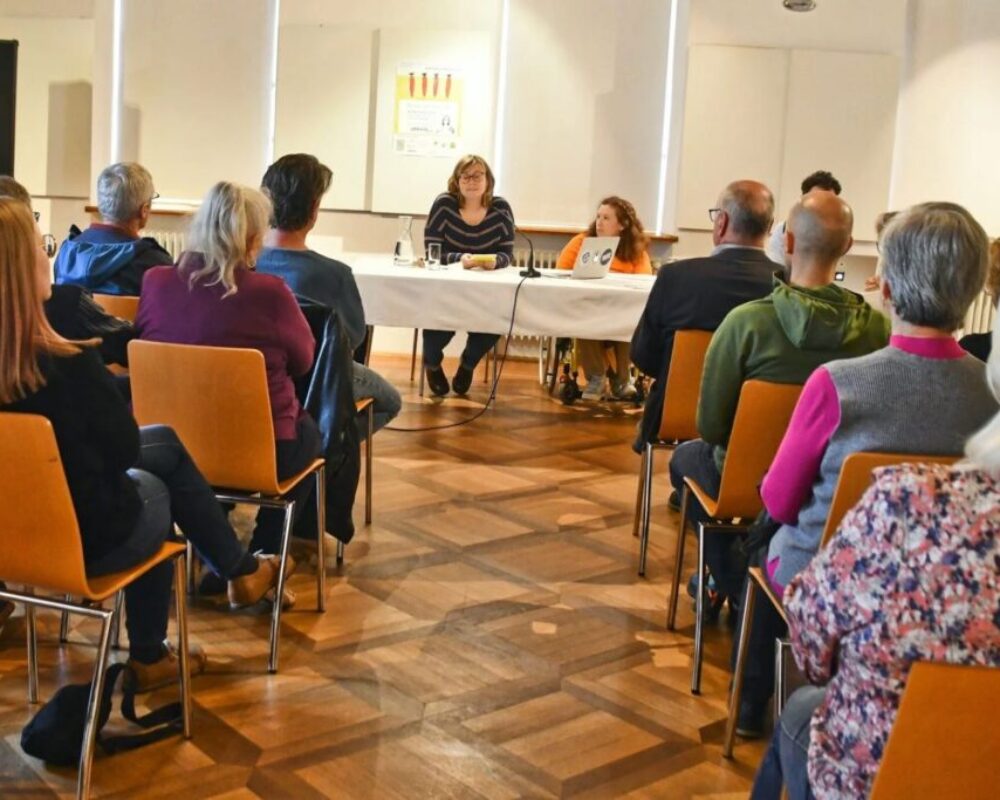 Ein Gang, von dem aus links und rechts Menschen sitzen und zuhören. Am Ende des Ganges sitzen links Hannah Wahl und rechts Julia Hofmarcher an einem Tisch.
