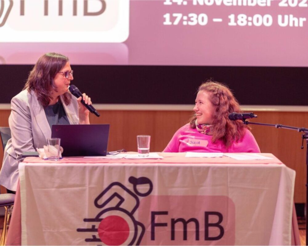 Heidemarie Egger sitzt links neben Julia und spricht in ein Mikrophon. Julia lächelt ihr zu.