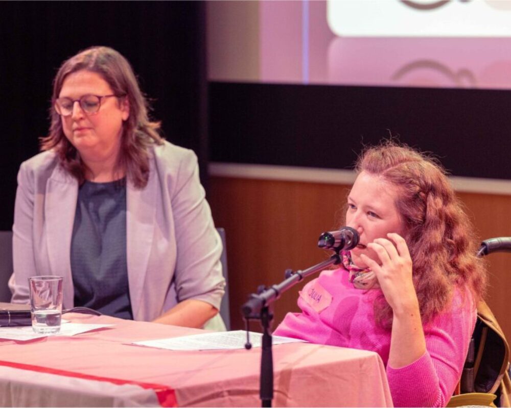 Heidi Egger sitzt links neben Julia, hört ihr zu. Julia spricht ins Mikro und hebt dabei die Linke Hand.