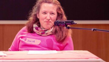 Julia sitzt hinter einem Tisch, lächelt mit einem Mikrophon vor dem Mund