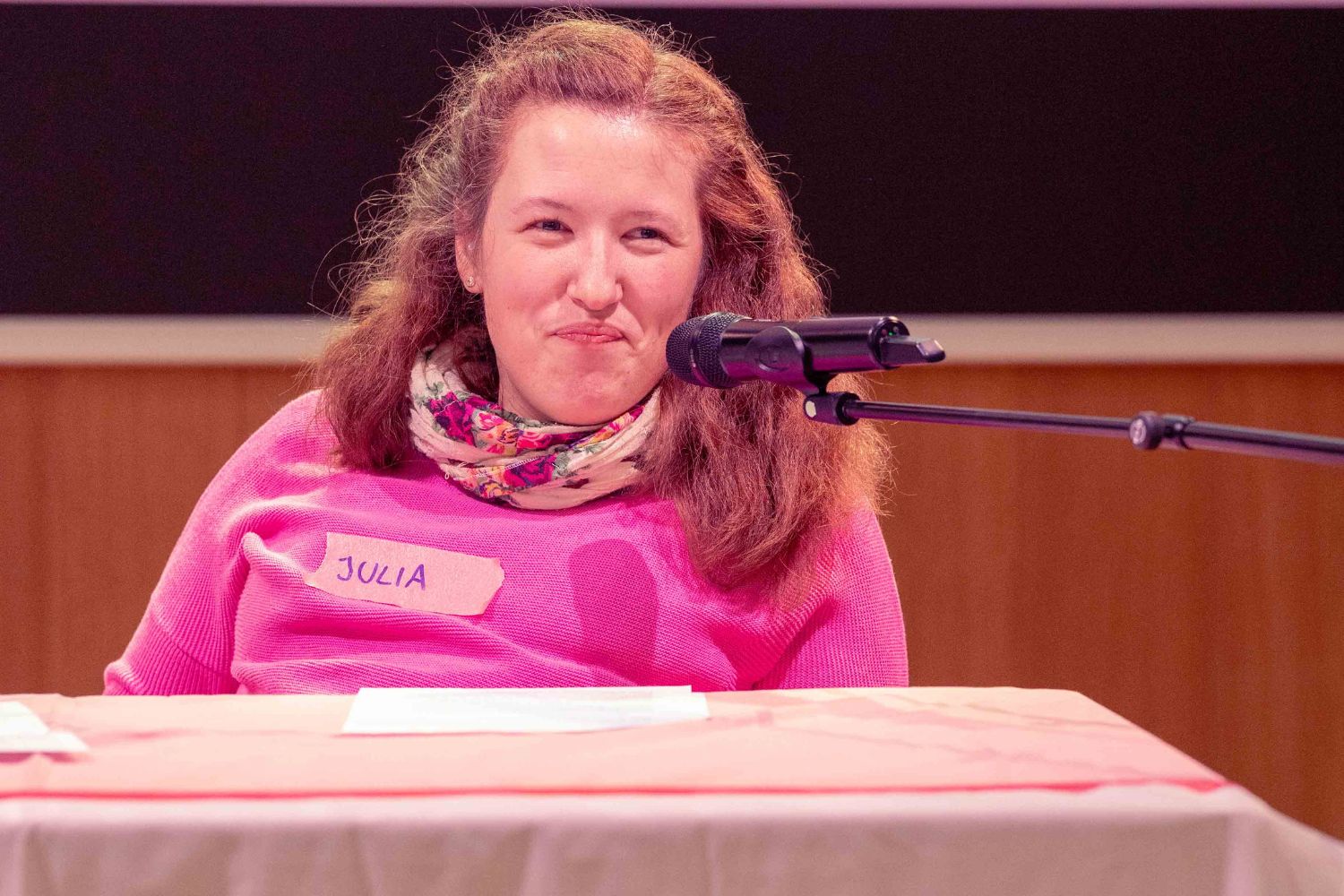 Julia sitzt hinter einem Tisch, lächelt mit einem Mikrophon vor dem Mund